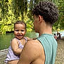 baby, child, young_adult, river, water, trees, greenery, smile, happy, outdoor, nature, holding, person, sleeveless_shirt, curly_hair, grass, people, summer, park, daytime