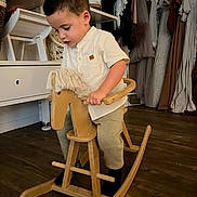 Matis participe au concours pour gagner de l'argent avec cette photo : child, toddler, rocking_horse, wooden_toy, indoor, clothing, floor, furniture, curly_hair, white_shirt, pants, black_socks, shelf, hanger, dress, home_decor, wood_floor, play, boy, concentration