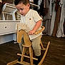 child, toddler, rocking_horse, wooden_toy, indoor, clothing, floor, furniture, curly_hair, white_shirt, pants, black_socks, shelf, hanger, dress, home_decor, wood_floor, play, boy, concentration