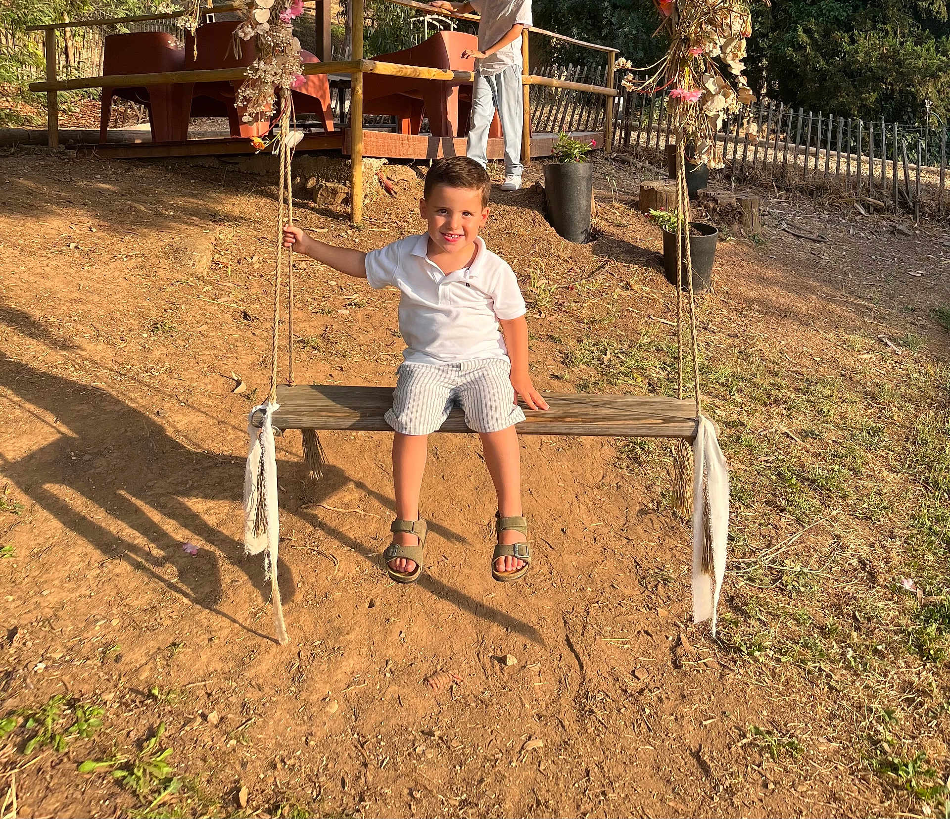 Matis participe au concours pour gagner de l'argent avec cette photo : child, boy, swing, outdoor, sunlight, dirt, wooden_swing, rope, smiling, casual_clothing, shorts, sandals, deck, plant_pots, fence, nature, daylight, happy, play, recreation