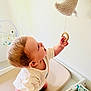 baby, child, curious, indoor, toy, moon, changing_table, nursery, hand, skin, clothing, white_wall, wooden_ring, soft_toy, infant, person, room, baby_girl_or_boy, looking_up, holding