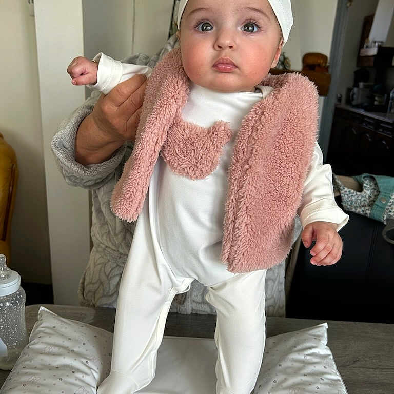 Tyssiana a rejoint le concours — aidez-le/la à gagner de superbes lots ! baby, baby_bottle, child, clothing, curious, cushion, face, hand, hat, home, indoor, infant, onesie, person, pink_vest, pom_poms, soft_lighting, standing, support, table