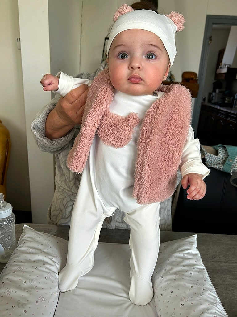 Tyssiana a rejoint le concours — aidez-le/la à gagner de superbes lots ! baby, infant, child, onesie, pink_vest, hat, pom_poms, hand, support, indoor, table, baby_bottle, cushion, face, person, clothing, curious, standing, soft_lighting, home