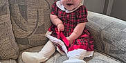 Amelia is registered to the contest to win money with this photo: baby, child, infant, smile, red_bow, dress, plaid, couch, furniture, white_tights, footwear, headband, happy, indoors, person, cute, patterned_fabric, seated, small_child, cozy