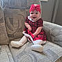 baby, child, infant, smile, red_bow, dress, plaid, couch, furniture, white_tights, footwear, headband, happy, indoors, person, cute, patterned_fabric, seated, small_child, cozy