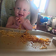 Sebastien is registered to the contest to win money with this photo: baby, blue_eyes, child, clothing, face, feeding, food, hair, happy, high_chair, indoor, kitchen, meal, messy, person, plate, smiling, spaghetti, tray, window