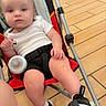 baby, child, stroller, white_shirt, black_shorts, black_sneakers, blanket, wooden_floor, indoor, person, footwear, seat, curious, young_child, casual_clothing, baby_bottle, floor_tiles, baby_face, toddler, relaxed
