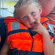 Nina a rejoint le concours — aidez-le/la à gagner de superbes lots ! child, life_jacket, orange, blue_eyes, blonde_hair, braids, playful, smiling, sitting, outdoor, summer, boat, safety, person, portrait, cute, casual_clothing, bright_colors, fun, relaxed