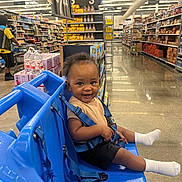 Janari is registered to the contest to win money with this photo: toddler, child, shopping_cart, grocery_store, aisle, smile, blue, seatbelt, floor, shopping, person, socks, clothing, retail, store, shelf, products, light, happy, indoor
