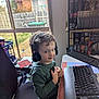 child, headphones, computer, keyboard, desk, chair, indoor, curly_hair, smile, sitting, window, backyard, board_games, shelf, toy, person, technology, room, casual, young