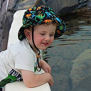 Oliver is registered to the contest to win money with this photo: child, toddler, hat, dinosaur_pattern, water, pool, rock, white_ledge, curious, outdoor, smiling, short_sleeve_shirt, blue_shorts, daylight, portrait, nature, playing, young, happy, casual_clothing