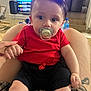 Cyriak participe au concours pour gagner de l'argent avec cette photo : baby, pacifier, red_shirt, black_shorts, person, hand, living_room, television, furniture, floor, tattoo, water_bottle, sitting, indoor, child, skin, clothing, face, eyes, holding_hand
