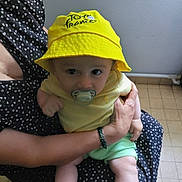 Cyriak participe au concours pour gagner de l'argent avec cette photo : baby, child, pacifier, yellow_hat, tour_de_france, adult_hand, indoor, tiled_floor, curious_expression, holding, black_dress, white_hearts, shorts, green_shorts, yellow_shirt, face, person, seated, wall, bracelet