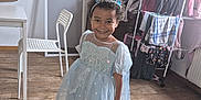 Maëlys a rejoint le concours — aidez-le/la à gagner de superbes lots ! child, smiling, princess_dress, tiara, barefoot, wooden_floor, indoor, clothing_rack, white_chair, table, curtain, wall_art, happy, person, portrait, sparkle, dress, floor, room, home