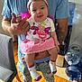 baby, child, infant, pink_skirt, headband, socks, adult, holding, indoor, water_dispenser, wooden_floor, blue_shirt, ripped_jeans, happy, playful, tongue_out, person, casual_clothing, home, furniture