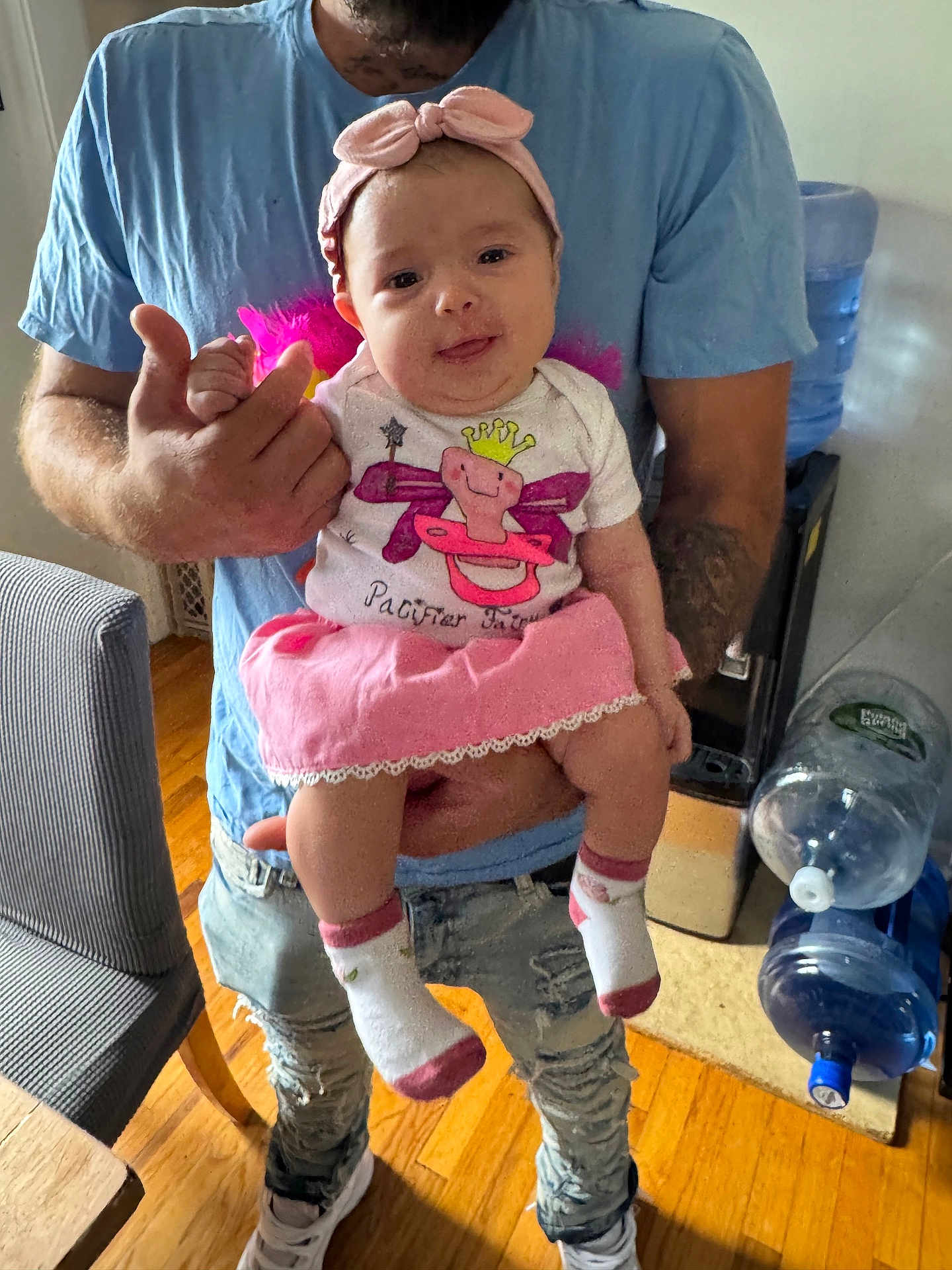 Jessenia joined the competition — help win amazing prizes! baby, child, infant, pink_skirt, headband, socks, adult, holding, indoor, water_dispenser, wooden_floor, blue_shirt, ripped_jeans, happy, playful, tongue_out, person, casual_clothing, home, furniture