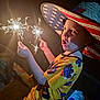 child, boy, hat, american_flag, sparkler, fireworks, night, yellow_clothing, expression, excitement, celebration, face, lighting, hand, sitting, indoors, party, fun, youth, portrait