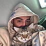 person, cat, hoodie, tabby_cat, face, eyes, furry, pet, cozy, indoor, snuggling, close_up, sleeping_cat, comfort, blurred_background, casual_clothing, animal, warm, cute, relaxed