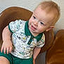 toddler, child, smiling, sitting, chair, leather_chair, shorts, shirt, patterned_clothing, indoor, light_skin, blond_hair, blue_eyes, happy, curious, portrait, young_child, casual, home, wall