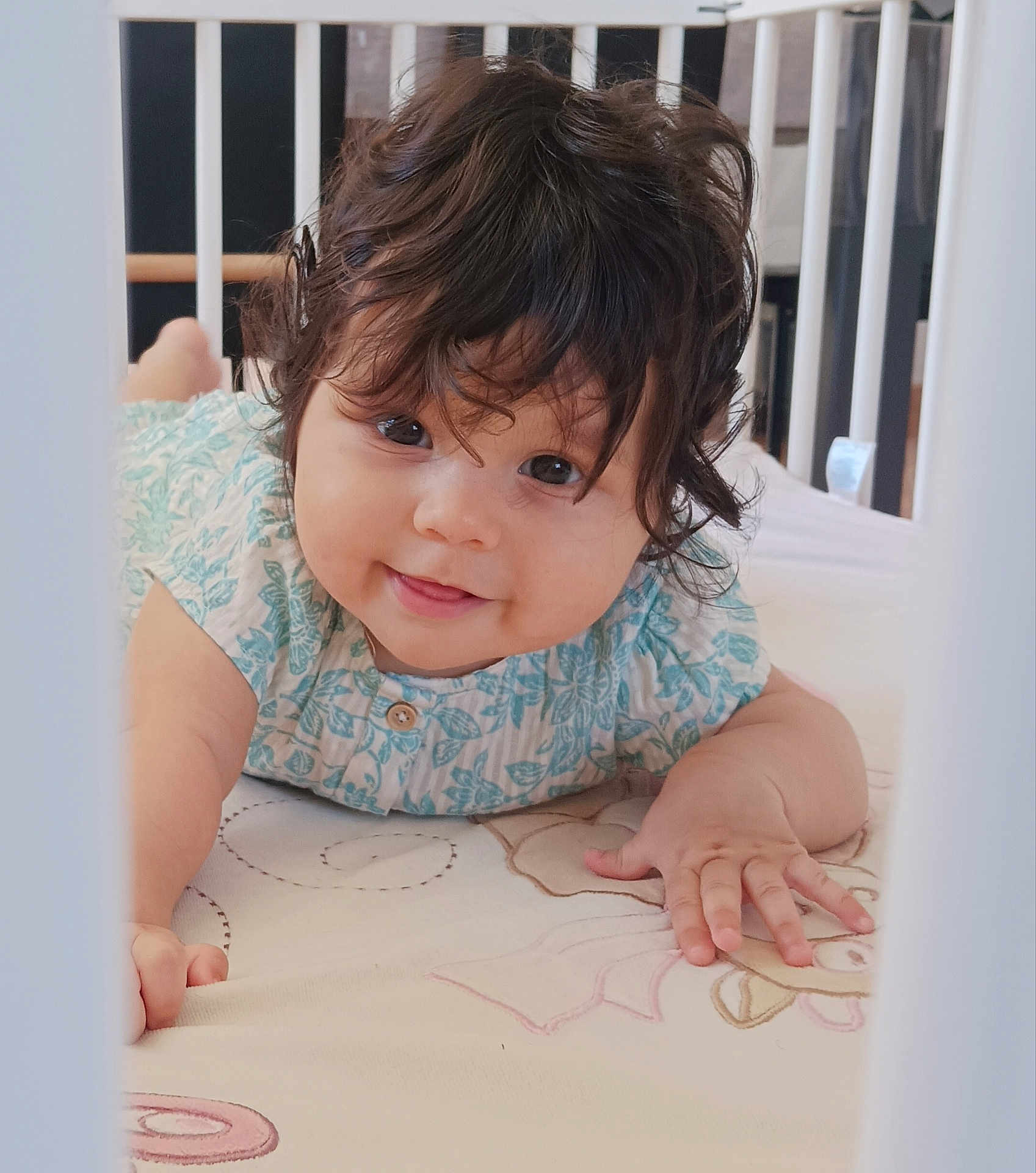 Catalina participe au concours pour gagner de l'argent avec cette photo : baby, child, crib, tummy, curly_hair, blue_clothing, smiling, hand, face, eyes, indoor, cute, infant, bedroom, fabric, pattern, skin, person, young, happy