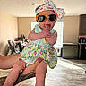Aria is registered to the contest to win money with this photo: baby, child, sunglasses, headband, floral_dress, tattoo, hands, indoor, window, flooring, kitchen, appliances, high_chair, person, cute, infant, holding, light, home, fashion