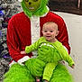 baby, child, person, costume, grinch, christmas, holiday, green_fur, santa_suit, tree, decorations, indoor, smiling, seated, shoes, red, white, green, festive, celebration
