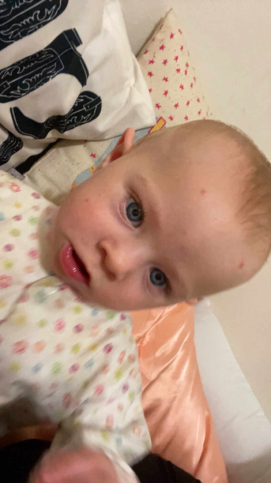 Aurora West is registered to the contest to win money with this photo: baby, child, face, blue_eyes, onesie, polka_dot, pillow, star_pattern, bed, blanket, indoor, closeup, infant, skin, head, person, cute, expression, young, sleepwear