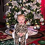 Aurora West is registered to the contest to win money with this photo: baby, child, christmas_tree, ornaments, silver_bows, leopard_print_onesie, white_chair, holiday_rugs, toy_train, indoor, festive, smiling, cute, decorations, lights, floor, holiday, seasonal, happy, portrait