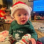 baby, child, santa_hat, christmas_pajamas, holiday, living_room, girl, presents, television, decorations, floor, carpet, table, chair, face, person, indoors, smiling, cozy, festive