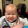 baby, child, smiling, food, bib, spoon, highchair, indoor, person, face, infant, cute, cheeky, baby_seat, patterned_clothing, happy, home, furniture, feeding, playful