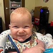 baby, baby_seat, bib, cheeky, child, cute, face, feeding, food, furniture, happy, highchair, home, indoor, infant, patterned_clothing, person, playful, smiling, spoon
