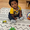 child, toddler, baby, indoor, rug, toy, yellow_shirt, curly_hair, socks, floor, wooden_floor, patterned_rug, looking_up, cute, expression, play, colorful, furniture, home, person