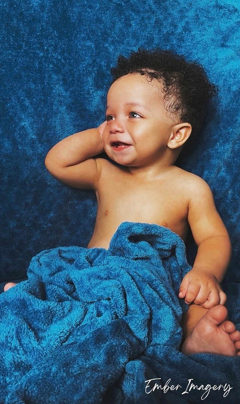 Vincent is registered to the contest to win money with this photo: baby, child, smile, curly_hair, blue_blanket, soft_texture, happy, portrait, infant, bare_chest, cute, relaxed, cozy, indoors, skin, hand, foot, face, expression, photography