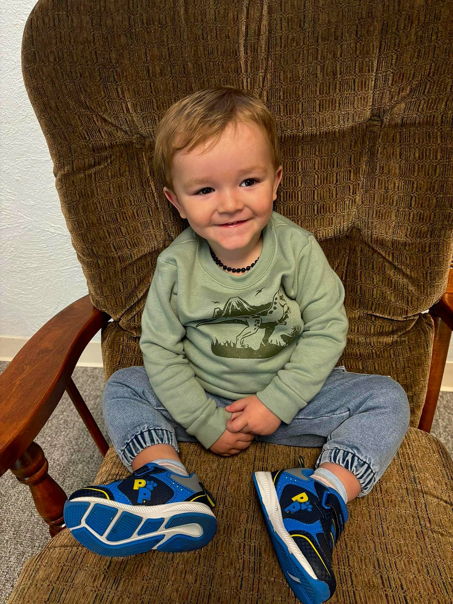 Lucas is registered to the contest to win money with this photo: baby_toddler_clothing, chair, cheek, comfort, eye, face, flooring, foot, hairstyle, happy, human_leg, joy, knee, leg, person, sitting, sleeve, smile, sneakers, standing