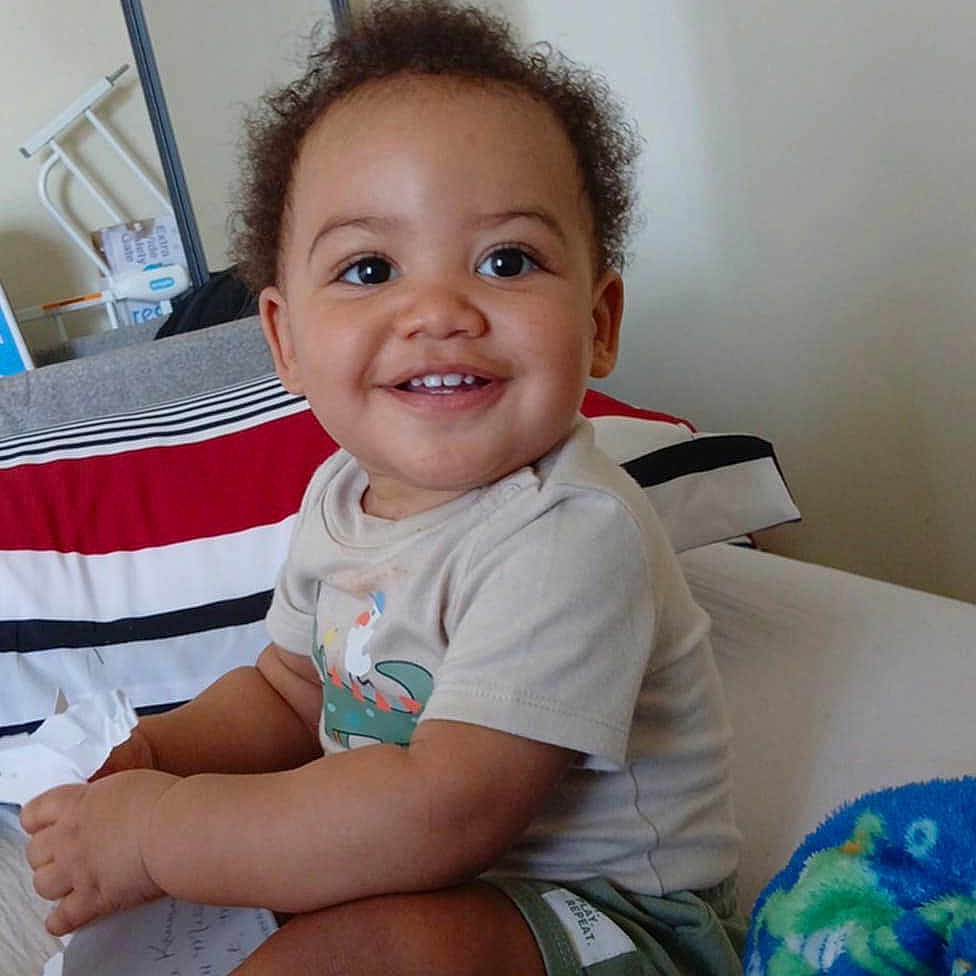 Asher is registered to the contest to win money with this photo: bed, blanket, bright, child, clothing, cozy, curly_hair, furniture, happy, home, indoor, mirror, paper, person, portrait, shorts, sitting, smiling, toddler, tshirt