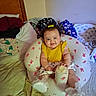 baby, infant, child, smiling, sitting, portrait, bedroom, bed, blanket, pillow, clothing, yellow_shirt, hair_bow, bottle, baby_seat, bunny_pattern, indoor, cozy, happy, face