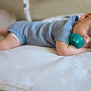 Wesley is registered to the contest to win money with this photo: baby, sleeping, pacifier, blue_onesie, infant, bed, white_bedspread, peaceful, cute, newborn, child, resting, indoors, soft_light, portrait, small_feet, head, face, hand, relaxation