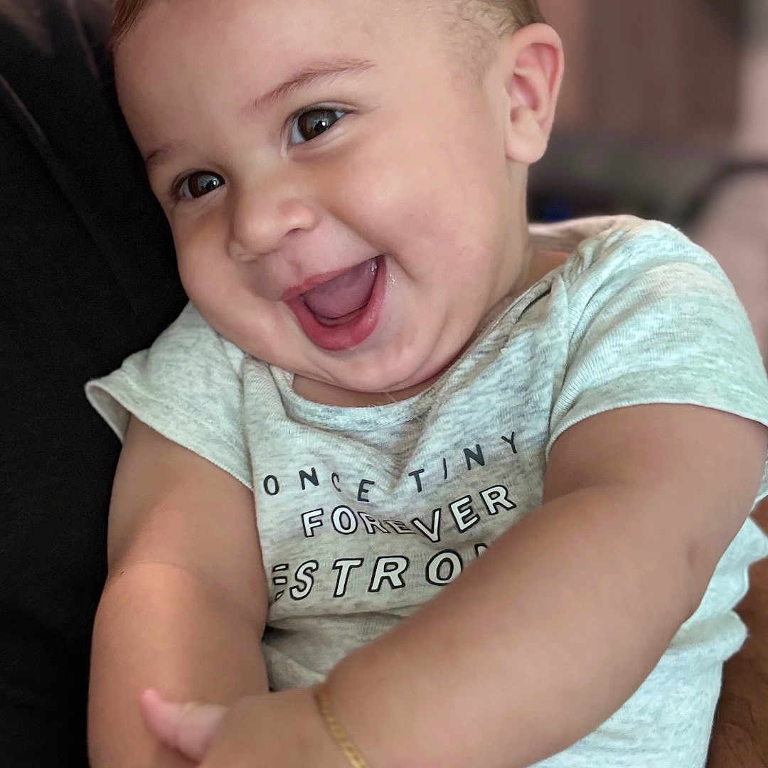Matteo Fernandes is registered to the contest to win money with this photo: arm, baby, blurred_background, bracelet, child, closeup, cute, expression, face, hands, happy, holding, human, indoor, infant, person, shirt, skin, smiling, young