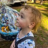 toddler, child, outdoor, forest, gift_basket, wrapped_gift, picnic_table, curly_hair, side_profile, nature, sunlight, clothing, patterned_shirt, greenery, trees, daylight, expression, looking_up, cute, young_child
