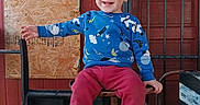 Domenick is registered to the contest to win money with this photo: toddler, child, smiling, blue_sweater, red_pants, sneakers, chair, metal, wooden_wall, boarded_window, outdoor, happy, portrait, person, hair, playful, casual_clothing, daylight, sitting, cute