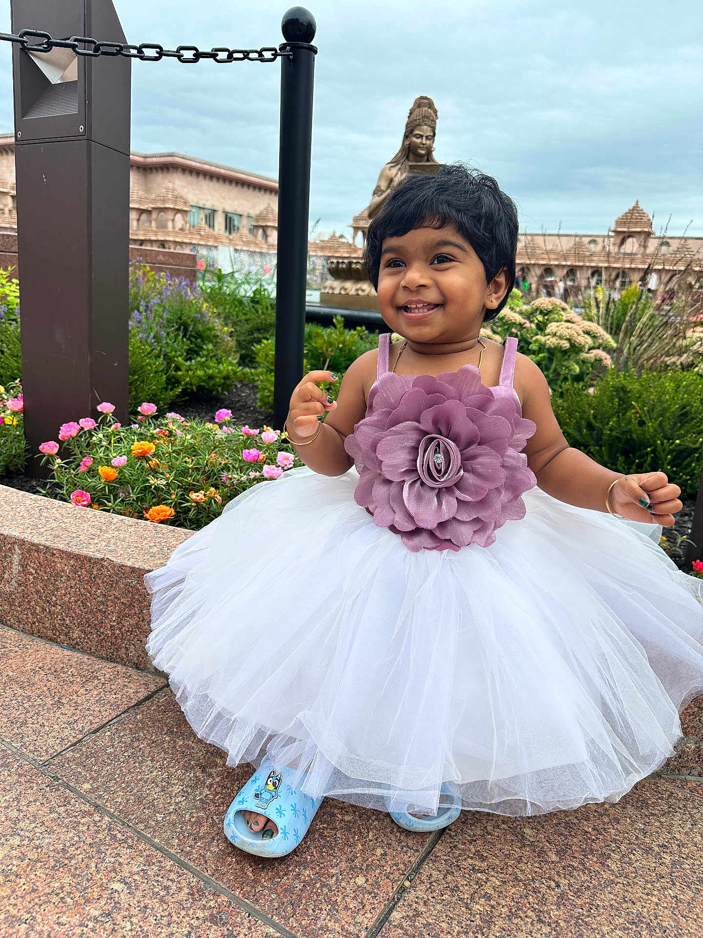 Diya is registered to the contest to win money with this photo: child, toddler, smiling, dress, flower, outdoor, garden, plants, statue, architecture, cloudy_sky, stone_pavement, happy, person, footwear, blue_slippers, nature, greenery, portrait, cute