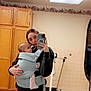 baby, baby_carrier, bathroom, black_sweatshirt, casual_clothing, ceiling_light, child, holding_baby, home_environment, indoor, mirror_selfie, mobile_phone, person, playful_expression, red_pants, reflection, scale, tiled_floor, woman, wooden_cabinets