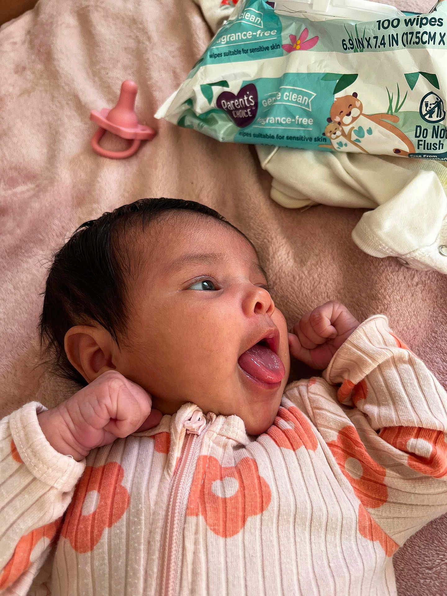 Kariah is registered to the contest to win money with this photo: baby, baby_wipes, blanket, child, clothing, cute, expression, face, floral_pattern, indoor, infant, lying_down, newborn, onesie, pacifier, pink, playful, small_hands, soft_texture, tongue_out