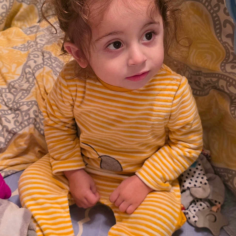 Brando joined the competition — help win amazing prizes! baby_bottle, bed, blanket, child, cozy, curious_expression, curly_hair, cute, face, home, indoors, infant, person, sitting, soft_lighting, striped_clothing, toddler, toy, yellow_onesie, young_child