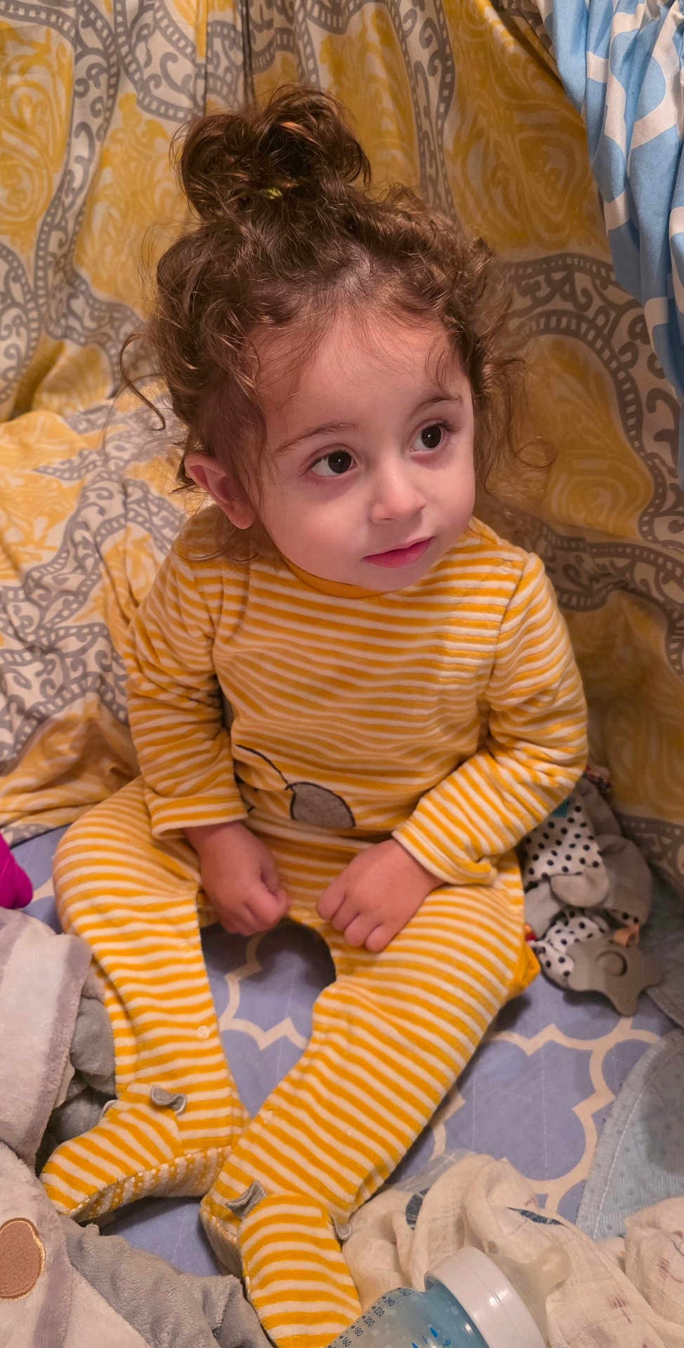 Brando joined the competition — help win amazing prizes! toddler, child, curly_hair, yellow_onesie, striped_clothing, sitting, indoors, bed, blanket, toy, baby_bottle, cute, soft_lighting, curious_expression, face, person, cozy, home, infant, young_child