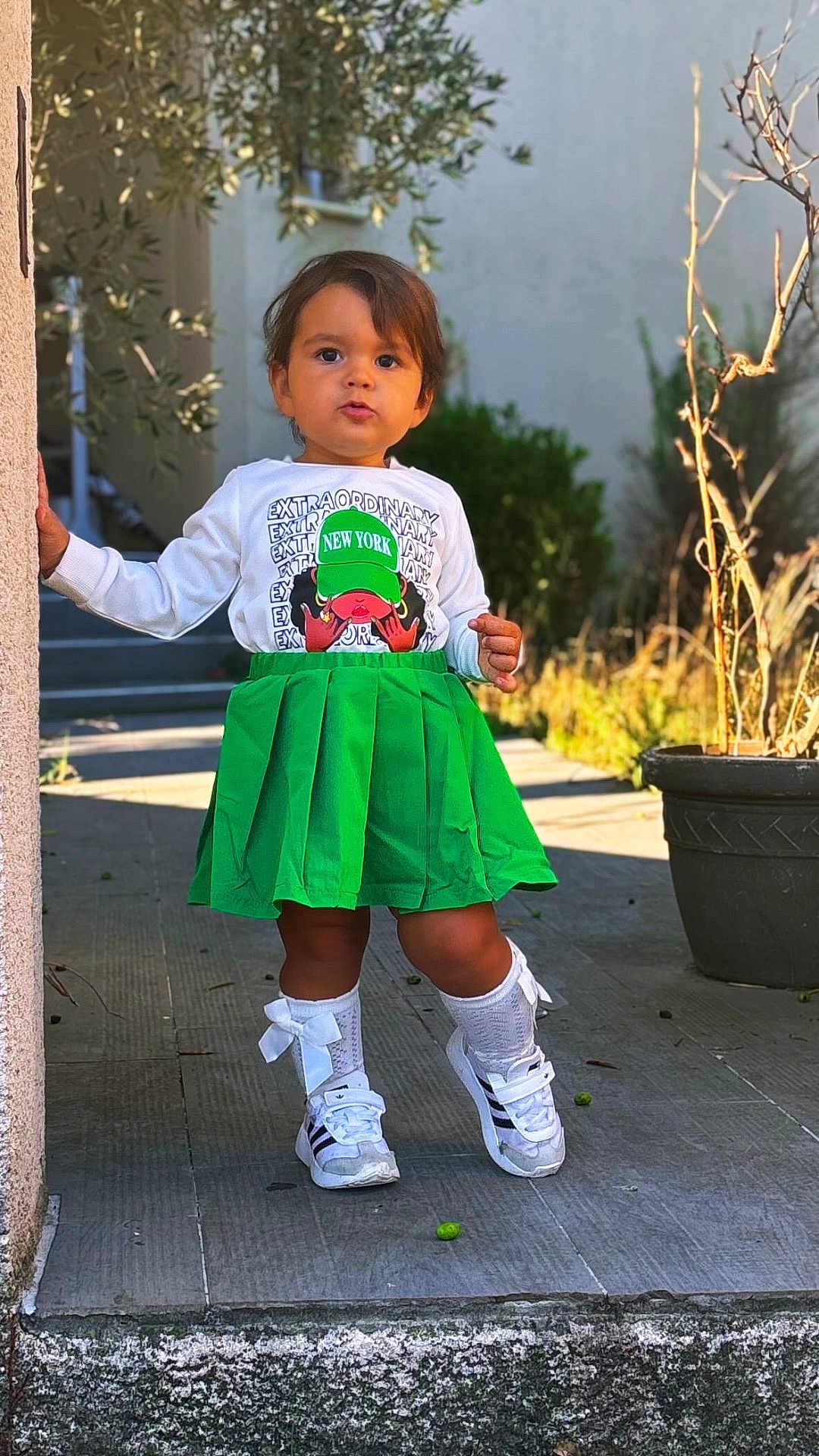 Alba participe au concours pour gagner de l'argent avec cette photo : toddler, child, green_skirt, white_sneakers, bows, outdoor, sidewalk, plant_pot, sunlight, wall, building, person, standing, young_child, casual_clothing, nature, daylight, expression, hair, portrait