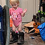 Maria is registered to the contest to win money with this photo: toddler, child, boots, pink_onesie, indoor, door, vacuum_cleaner, floor, clutter, brown_boots, cardboard_box, water_bottle, clothing, person, standing, curly_hair, light_hair, home, messy_room, looking_down