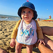 Loéna a rejoint le concours — aidez-le/la à gagner de superbes lots ! toddler, child, beach, sand, ocean, hat, bib, sunlight, shadow, water, people, summer, vacation, legs, hands, waves, sky, outdoor, daytime, casual