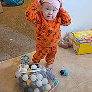 Vylette is registered to the contest to win money with this photo: toddler, child, indoor, pajamas, orange, pink_hat, tiles, toys, balls, mesh_bag, floor, standing, curious, expression, clutter, box, carpet, pacifier, baby, person