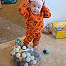 toddler, child, indoor, pajamas, orange, pink_hat, tiles, toys, balls, mesh_bag, floor, standing, curious, expression, clutter, box, carpet, pacifier, baby, person